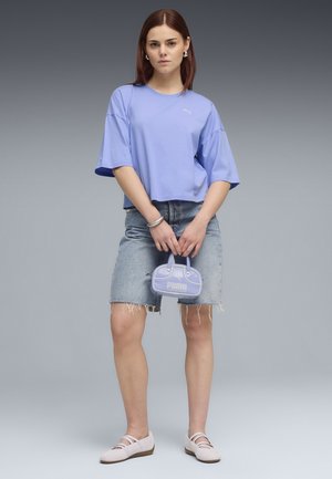 Jonge vrouw staand, draagt een losse lavendelkleurige T-shirt, een gescheurde denim rok, lichte schoenen en houdt een kleine lavendelkleurige Puma-handtas vast.