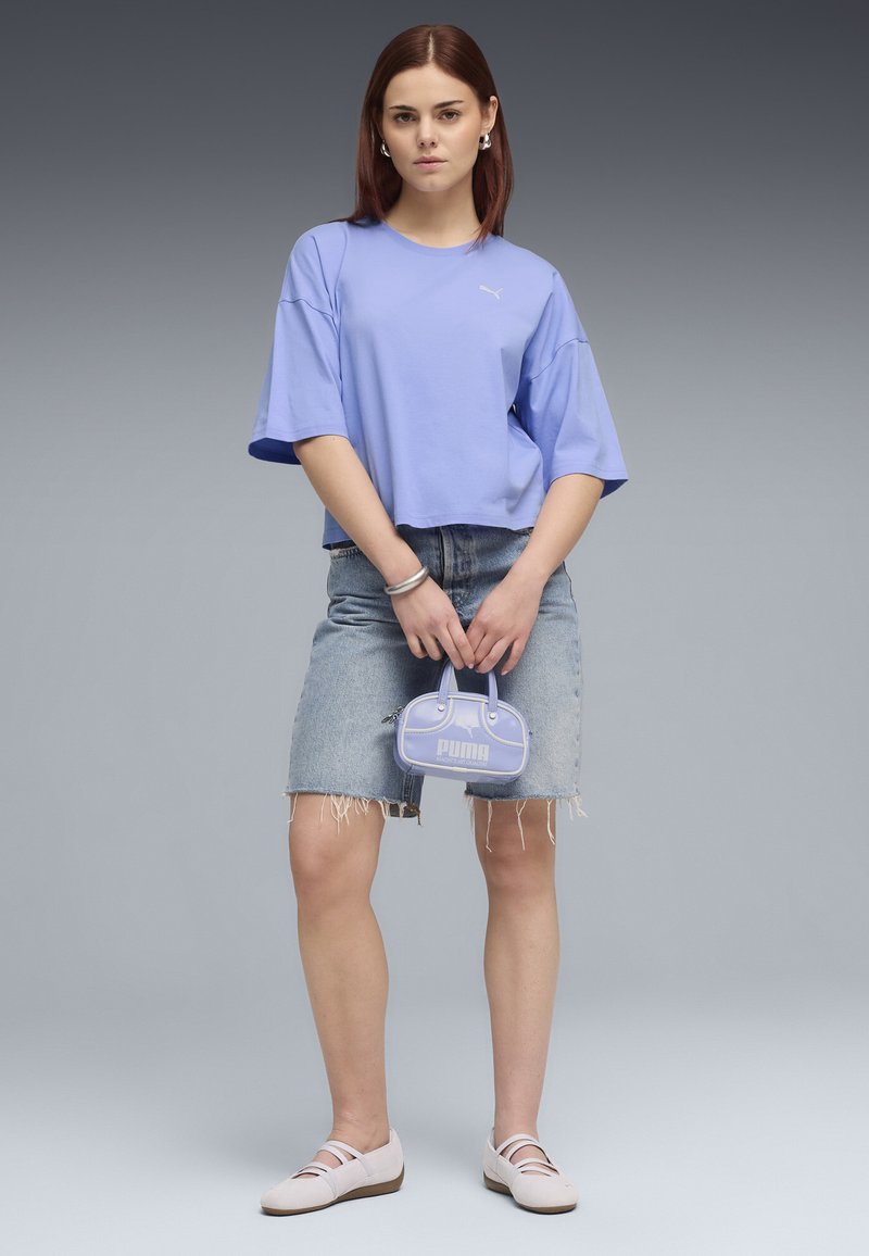 Jeune femme debout portant un T-shirt lavande ample, une jupe en jean effilochée, des chaussures claires, tenant un petit sac à main Puma lavande.
