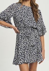 Kukallinen mekko, jossa on tummansininen tausta, lyhyet röyhelösateet, elastinen vyötärö ja väljä malli. Valkeat kukat luovat kontrastista yksityiskohtaa.