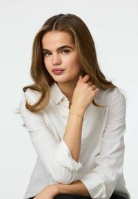 Modelka ubrana w białą koszulę z kołnierzykiem, z długimi, falującymi brązowymi włosami, jasną cerą i subtelną srebrną bransoletką na nadgarstku. Ręce ułożone delikatnie.