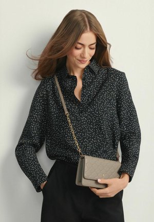 Kobieta ubrana w czarną bluzkę z długim rękawem w białe kropki, czarne spodnie, trzyma szarą pikowaną torebkę na ramię z złotym łańcuszkiem.