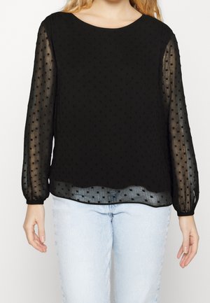 Vrouw draagt een zwarte, doorschijnende blouse met lange mouwen en polkadots, en lichtblauwe spijkerbroek, staand tegen een witte achtergrond.