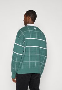 Πράσινο πλεκτό πουλόβερ με σχέδιο πλέγματος, λευκές ρίγες και λευκό γιακά. Διαθέτει λογότυπο στην πλάτη. Χαλαρή εφαρμογή και υφασμένο ύφασμα.