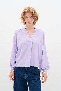 Blusa lavanda con scollo a V e polsini arricciati. Realizzata in un tessuto leggero, presenta un design morbido con una piega frontale, abbinata a jeans blu.