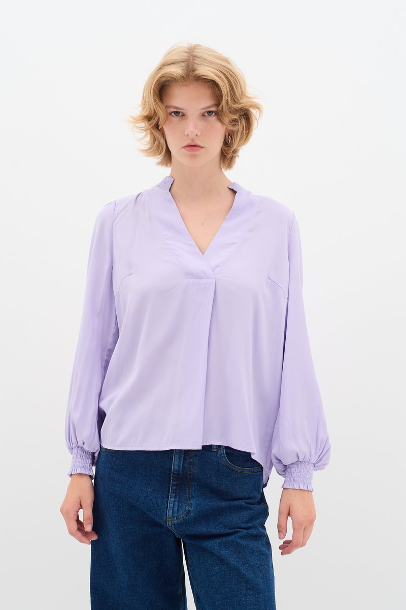 Blusa lavanda con scollo a V e polsini arricciati. Realizzata in un tessuto leggero, presenta un design morbido con una piega frontale, abbinata a jeans blu.