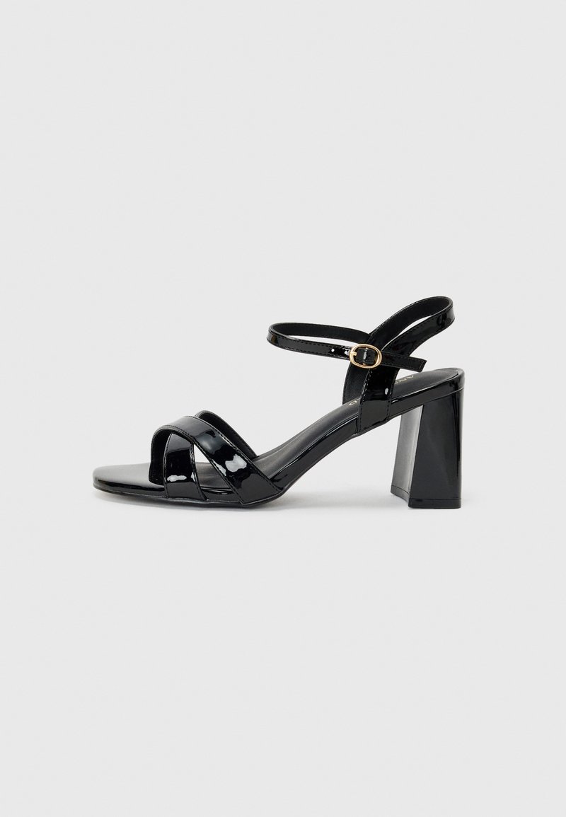 Black patent leather high-heel sandals with a block heel, featuring crisscross front straps and an adjustable ankle strap with a gold buckle.