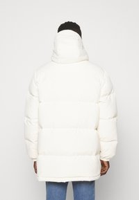 Muž v bílé péřové bundě s vysokým límcem a kapucí, viděn z boku na jednolitém bílém pozadí.