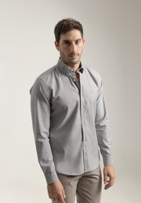 Camisa de botones gris con cuello, hecha de una tela texturizada. Presenta botones frontales y mangas largas, combinada con pantalones de color crema.