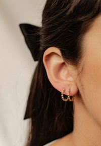 Złote kolczyki w formie obręczy z kręconym wzorem, zdobione małymi, okrągłymi koralikami, prezentowane na uchu modelki. Widoczne ciemne włosy i wstążka.