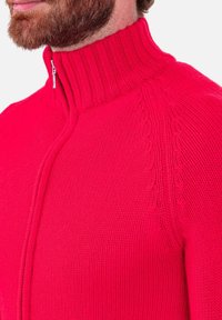 Pull à fermeture éclair ajusté de couleur rose vif. Présente un col côtelé et des détails texturés le long des épaules. Le matériau semble tricoté avec une finition lisse.