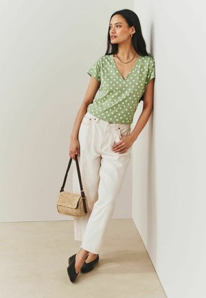 Woman leaning against white wall wearing green polka dot top, white pants, black shoes, holding woven handbag with dark straps.