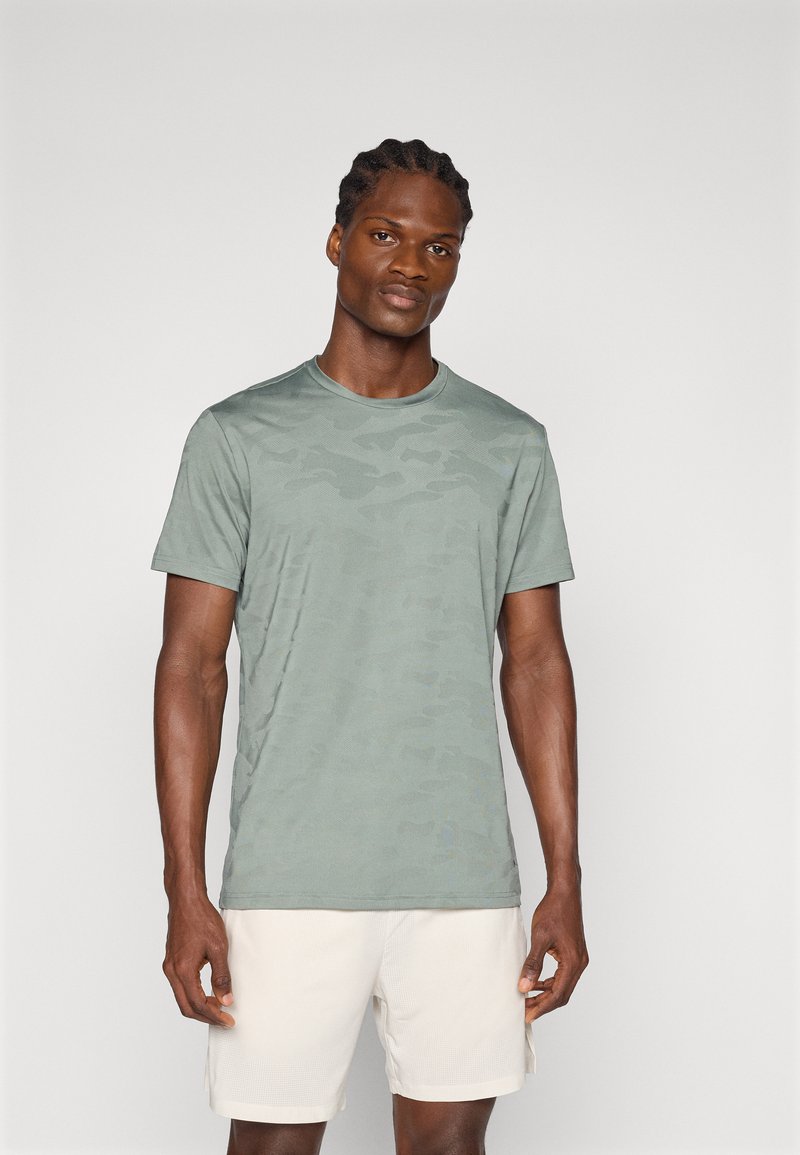 Camiseta de patrón de camuflaje verde claro, hecha de tela suave, con un cuello redondo estándar y mangas cortas, combinada con pantalones cortos beige.
