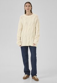 Pull en maille torsadée crème avec une coupe oversize, un col rond et des manches longues. Associé à un jean bleu foncé et des baskets marron.