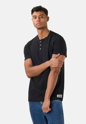 Giovane uomo con maglia henley nera a maniche corte e jeans blu, in piedi con un braccio che tiene l'altro su uno sfondo chiaro e uniforme.