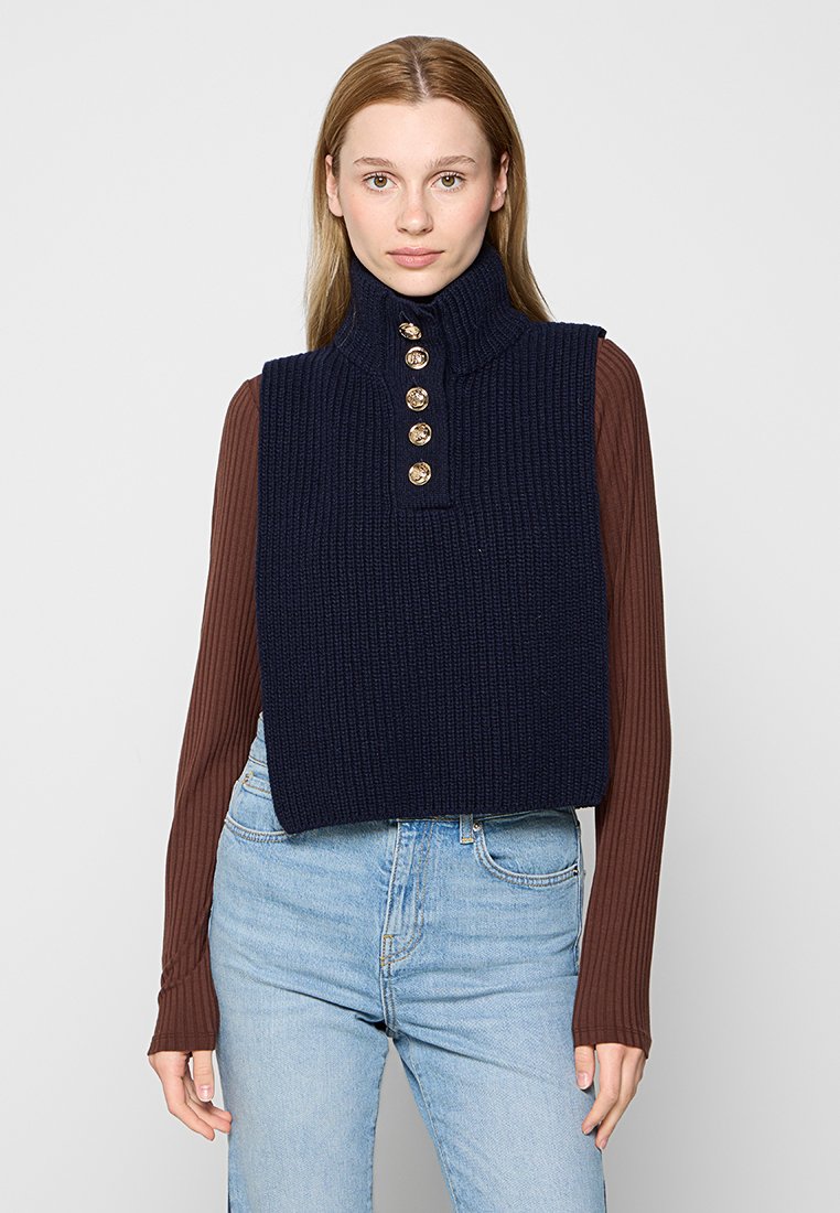 Haut sans manches en tricot marine avec un col montant et cinq boutons dorés, associé à une chemise brun à manches longues et un jean en denim bleu clair.