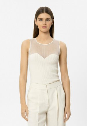 Junge Frau mit glattem Haar, trägt ein ärmelloses, weißes, geripptes Oberteil mit durchscheinendem oberen Einsatz und weiße, hoch taillierte Hose, steht vor einem einfarbigen Hintergrund.