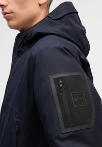 Námornícka bunda s kapucňou z textúrovaného materiálu, so čiernym nášivkom na ľavom rukáve s otvorenou kapsou a logom.