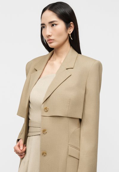 Γυναίκα με μακριά σκούρα μαλλιά, ντυμένη με μπεζ φόρεμα χωρίς τιράντες και υπερμεγέθη μπεζ σακάκι με βάτες στους ώμους, στέκεται μπροστά σε απλό φόντο.