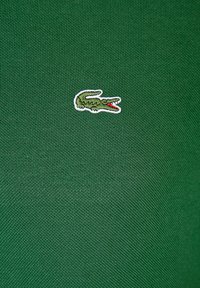 Žalia tekstūruota medžiaga su mažuoju išsiuvinėtu krokodilo logotipu raudonomis detalėmis, esančiu apatiniame dešiniajame kampe.