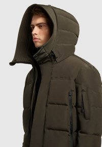 Pūkinė alyvuogių žalios spalvos striukė su aukštu apykakle ir dideliu, struktūruotu gobtuvu. Yra užtrauktukais užsegamos šoninės kišenės ir kontrastingos siūlės.