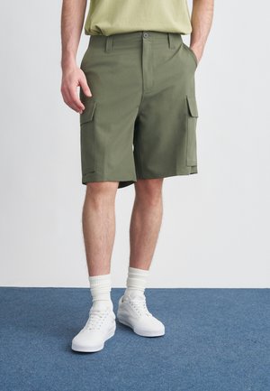 Olive rohelised vabaajajalanõud taskutega ees ja külgedel, nööbitava kinnituse ja voltidega, kombineeritud valgete tossude ja soonikkoes sokkidega.