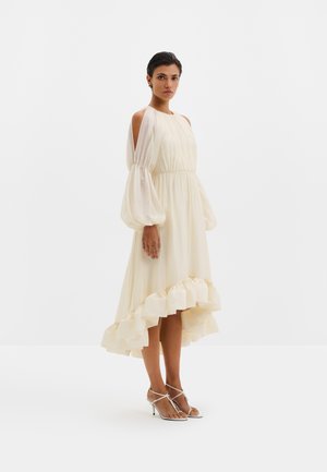 Femme portant une robe crème à manches longues bouffantes avec épaules découpées, ourlet à volants, et des sandales blanches à talons avec lanières.