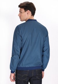 Giacca blu con un motivo texturizzato, colletto e polsini a coste, e vita blu uniforme. Indossata con jeans in denim scuro.