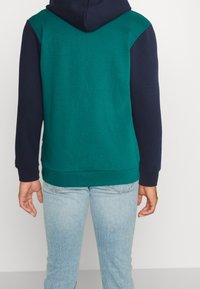 Sudadera con capucha de cuerpo verde y mangas azul marino. Presenta un bolsillo tipo canguro y puños de canalé. Fabricada con una suave mezcla de algodón.