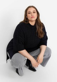 Mörk marinblå kabelstickad oversized tröja med ribbat knäbånd, i kombination med grå vida sweatpants och svarta klackboots vid ankeln.