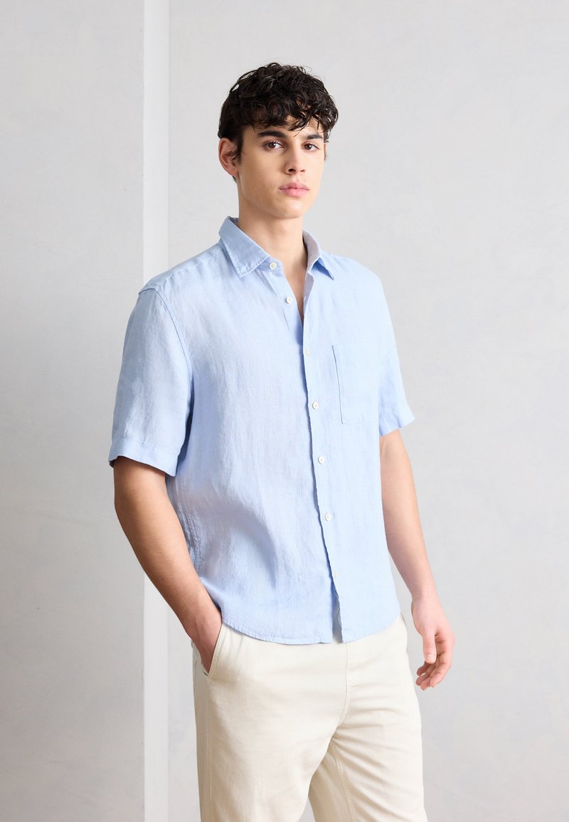 Νέος άνδρας που στέκεται με το ένα χέρι στην τσέπη, φορώντας γαλάζιο πουκάμισο με κοντά μανίκια και μπεζ παντελόνι, μπροστά σε απλό φόντο.