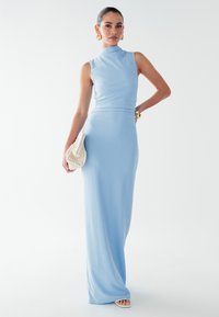 Světlomodré šaty bez rukávů s vysokým výstřihem. Přiléhavý korzet přechází do dlouhé rovné sukně. Kombinováno s bílou texturovanou kabelkou.