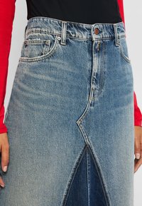 Høy midje lys blå denimskjørt med frontslitt, brukt med en svart topp og røde lange ermer, med hendene avslappet ved siden av.