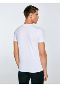 Fiatal férfi egyszínű fehér, rövid ujjú pólóban és fekete farmerben, hátulról nézve, feje jobbra fordítva, világos háttér előtt.