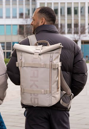 Bărbat purtând o geacă matlasată neagră, cu un rucsac bej mare cu design roll-top și curele, mergând în aer liber lângă o clădire modernă.