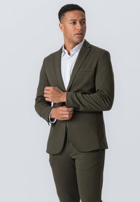 Olivengrønt jakkesæt med skræddersyet pasform, der har en enkeltknapjakke, notch-lapel og en brystlomme. Kombineret med en hvid skjorte.