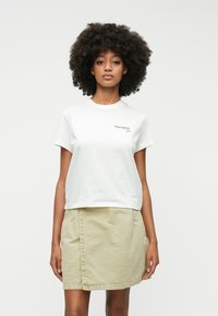 Camiseta blanca de algodón de manga corta, con un pequeño logo negro en el lado izquierdo del pecho. Combinada con una falda de tubo beige claro que llega a la altura de la rodilla.