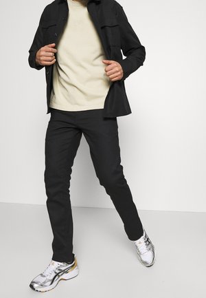 Homme portant une veste noire, un t-shirt beige, un pantalon noir et des chaussures de sport blanches, debout devant un fond clair uni.