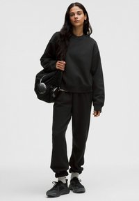 Schwarzer Sweatshirt und Sweatpants aus weichem Material; oversized Schnitt mit gerippten Bündchen. Getragen mit schwarzen Sneakers und einer schwarzen Tasche.