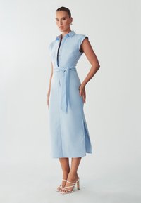 Himmelblaues, knielanges Hemdkleid mit kurzen Ärmeln, Kragen, Knopfleiste und Taillengürtel. Verfügt über seitliche Schlitze und ein glattes Material.