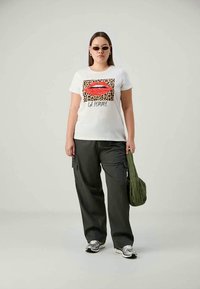 White t-shirt with red lips and leopard print background, paired with black cargo pants and gray sneakers. Model carries a green bag.