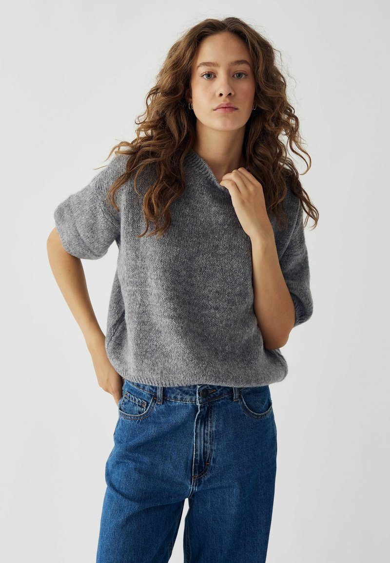 Maglione grigio lavorato a maglia con colletto alto e maniche corte, abbinato a jeans in denim blu a vita alta. Design semplice e testurizzato.