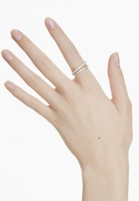 Drei ineinandergreifende Ringe aus Gold und Silber, mit einer Reihe kleiner klarer Steine auf einem Band. Glatte Oberfläche, getragen an einer Hand mit natürlichen Nägeln.
