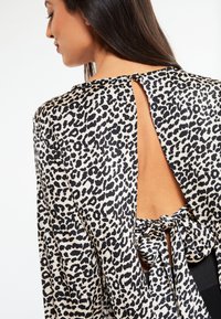 Bluse med leopardmønster, der har udskæring i ryggen og bindingslukning. Funktioner inkluderer blødt stof, flydende design og fremtrædende sort og creme mønster.