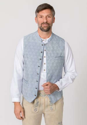 Mann trägt traditionelle bayerische Kleidung mit hellblauer gemusterter Weste, weißem Hemd und beigen bestickten Lederhosen.