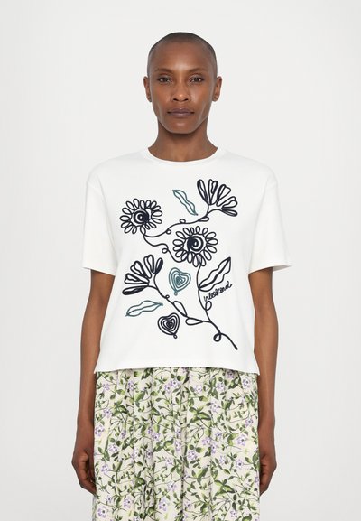 Weißes T-Shirt mit schwarzem und türkisfarbenem gesticktem Blumenmuster, kurze Ärmel, runder Hals, getragen mit einem Rock im Blumenmuster.