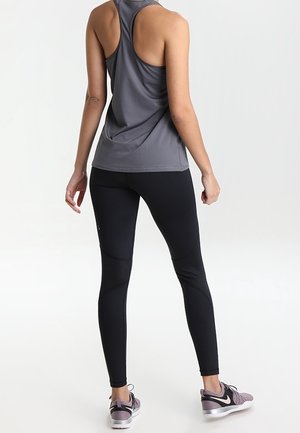 Personne portant un débardeur gris à dos nageur, des leggings noirs avec des panneaux en maille, et des baskets Nike grises, debout devant un fond blanc uni.