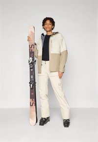 Άτομο με μπεζ και κρεμ σκι στυλ κρατώντας ένα όρθιο σκι, φορώντας μαύρες μπότες σκι, στέκεται μπροστά σε απλό ανοιχτό φόντο.