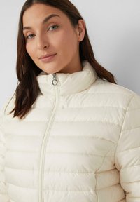 Cremfarbener Puffer-Jacke mit horizontalen Nähten, hohem Kragen und Frontreißverschluss. Glattes, glänzendes Material, taillierte Passform.