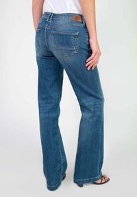 Jean flare en denim bleu présentant une taille mi-haute, deux poches arrière et une patch logo en cuir brun près de la taille. Texture lisse.