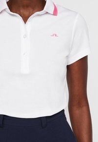 Kvinna som bär en vit kortärmad pikétröja med rosa kant på kragen och en liten rosa logotyp, i kombination med mörkblå byxor.